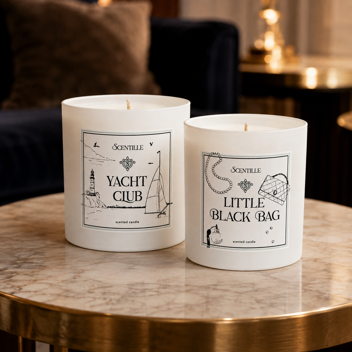 Two white scented candles by Scenttile, labeled 'Yacht Club' and 'Little Black Bag', displayed on a polished marble and brass table in a warm, upscale interior setting.