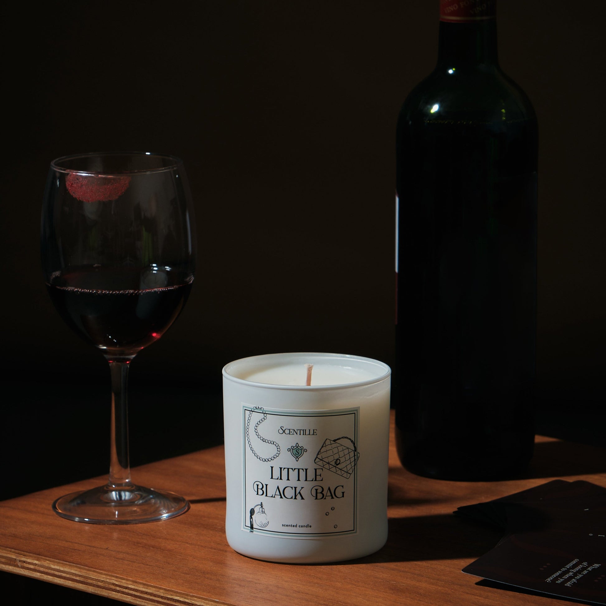 A Little Black Bag candle next to a glass of wine and a bottle